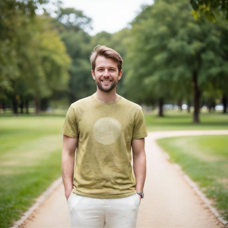 May include: A person wearing an olive green t-shirt with a faded white circle design. The person is also wearing white pants and a watch. The background is a park with a walking path and trees.