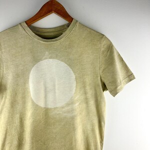 May include: A light olive green t-shirt with a large, off-white circle design on the front. The shirt is hanging on a wooden hanger against a white background. The t-shirt has short sleeves and a crew neck.