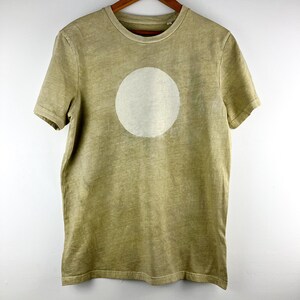 May include: A short-sleeved, olive green t-shirt with a large, off-white circle printed on the front. The shirt is hanging on a wooden hanger against a white background. The fabric has a slightly faded appearance.