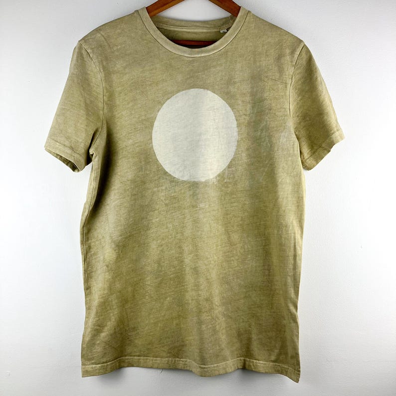 May include: A short-sleeved, olive green t-shirt with a large, off-white circle printed on the front. The shirt is hanging on a wooden hanger against a white background. The fabric has a slightly faded appearance.