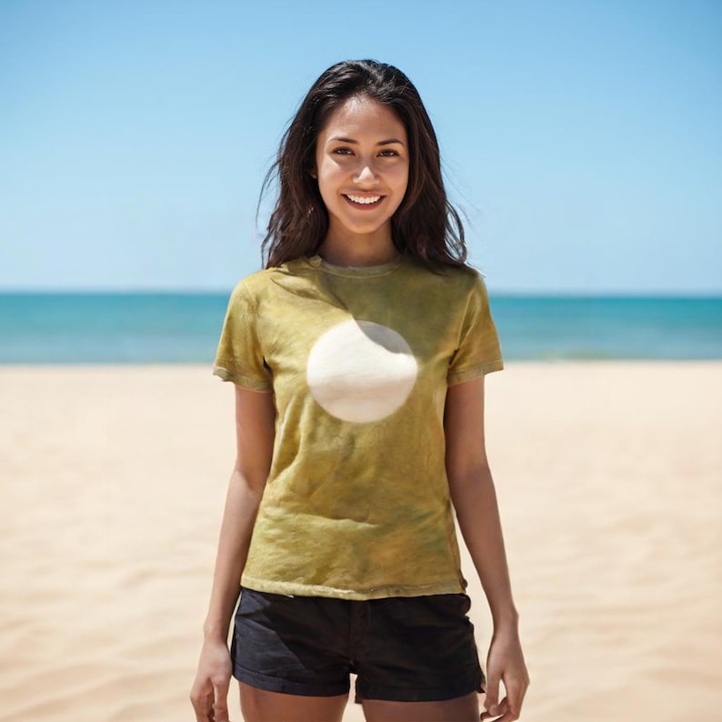 May include: A woman wearing a short-sleeved, olive-green t-shirt with a large white circle design. She is also wearing black shorts. The background shows a beach with sand, ocean, and a blue sky.