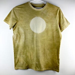 May include: A short-sleeved, olive green t-shirt with a large, off-white circle design on the front. The shirt has a crew neck and is hanging on a white hanger. The fabric has a slightly faded appearance.