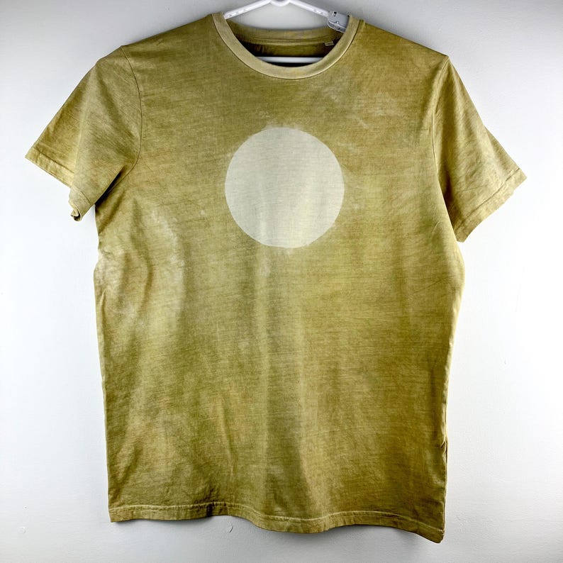 May include: A short-sleeved, olive green t-shirt with a large, off-white circle design on the front. The shirt has a crew neck and is hanging on a white hanger. The fabric has a slightly faded appearance.