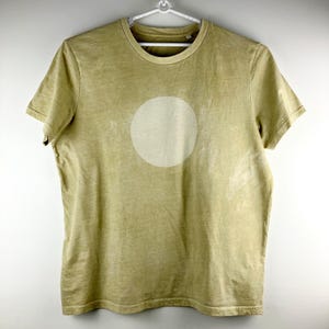 May include: A short-sleeved, olive green t-shirt with a large, off-white circle design on the front. The shirt is hanging on a white hanger against a white background. The fabric appears to have a slightly faded or vintage look.