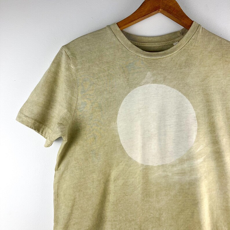 May include: A light olive green t-shirt with a large, off-white circle design on the front. The shirt has short sleeves and a crew neck. The fabric has a slightly faded appearance.