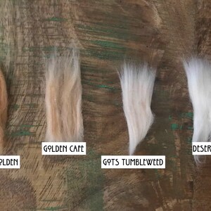 Op de afbeelding: Vier plukken vezels op een rij, gelabeld met witte rechthoekige kaartjes. De kleuren van links naar rechts zijn Deep Golden, Golden Cafe, Gots Tumbleweed en Desert Mist, met vari&euml;rende tinten van bruin, beige en wit.