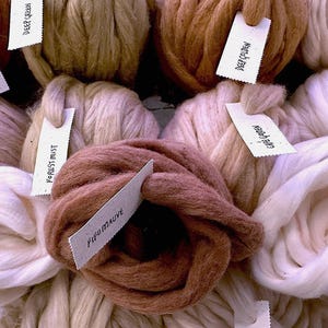 Op de afbeelding: Een verzameling dikke, grove bollen wol in diverse neutrale en aardse tinten, waaronder wit, cr&egrave;me, mauve en bruin. Elke bol is voorzien van een klein wit papieren label met handgeschreven teksten zoals Rich Cinnamon Choc, Deep Green, Deep Golden, Forest Mist en Fifo Mauve.