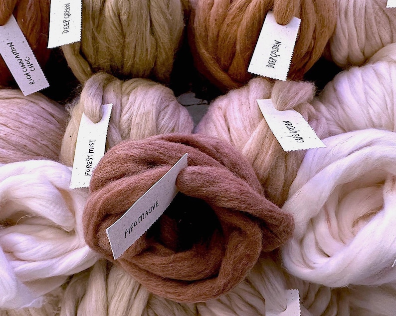 Op de afbeelding: Een verzameling dikke, grove bollen wol in diverse neutrale en aardse tinten, waaronder wit, cr&egrave;me, mauve en bruin. Elke bol is voorzien van een klein wit papieren label met handgeschreven teksten zoals Rich Cinnamon Choc, Deep Green, Deep Golden, Forest Mist en Fifo Mauve.