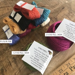 May include: Assortment of colorful yarn skeins in various sizes, including a 10 custom color mix, a 125 yard pull skein, and a 250 yard pull skein. The yarn is labeled "Ecobutterfly" and is made of organic Tangu cotton. The colors include magenta, wasabi, and a mix of other colors.