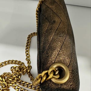 May include: A metallic gold and black shoulder bag with a chevron pattern. The bag features a gold chain strap and gold hardware. The bag is made of a textured material and has a flap closure.