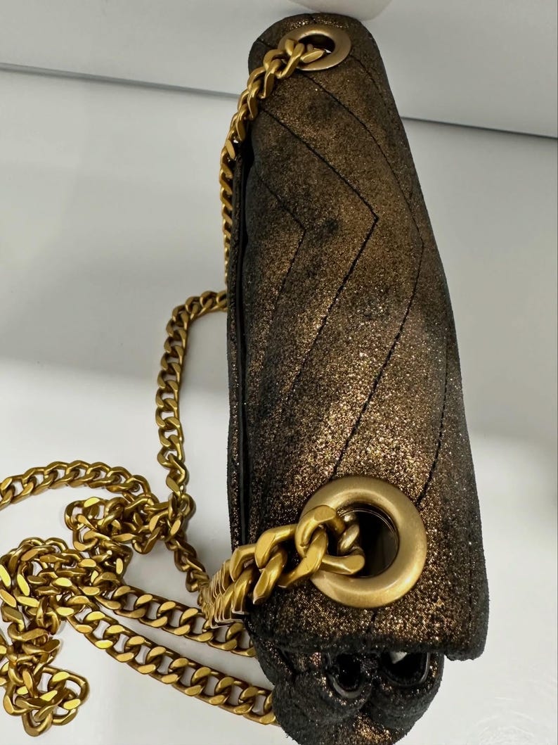 May include: A metallic gold and black shoulder bag with a chevron pattern. The bag features a gold chain strap and gold hardware. The bag is made of a textured material and has a flap closure.