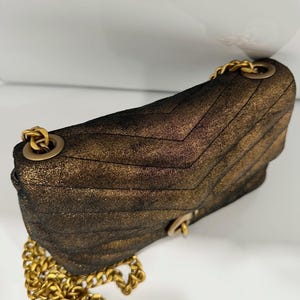 May include: A metallic bronze-colored shoulder bag with a quilted chevron pattern. The bag features a gold-tone chain strap and hardware. The bag is made of a textured material and has a flap closure.