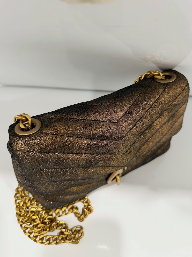 May include: A metallic bronze-colored shoulder bag with a quilted chevron pattern. The bag features a gold-tone chain strap and hardware. The bag is made of a textured material and has a flap closure.