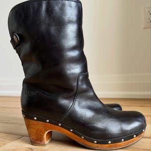 Puede incluir: Una bota de cuero negro a media pantorrilla con tacón y suela de madera, decorada con tachuelas metálicas plateadas a lo largo de la base. La bota incluye una correa lateral con un botón y el logotipo de la marca UGG grabado en el lateral.