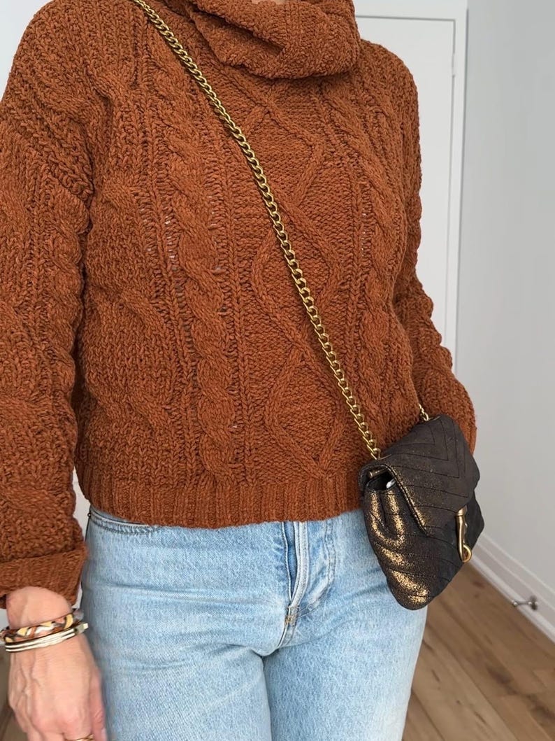 May include: A brown cable-knit sweater with a cowl neck, paired with light blue jeans. A small black and gold crossbody bag with a gold chain strap is worn. The person is also wearing bracelets.