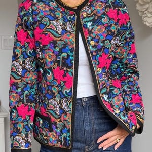May include: A long-sleeve quilted jacket featuring a vibrant floral and paisley pattern in shades of hot pink, royal blue, and teal on a black background. The jacket has a round neckline, black trim, and gold-toned buttons along the front closure.