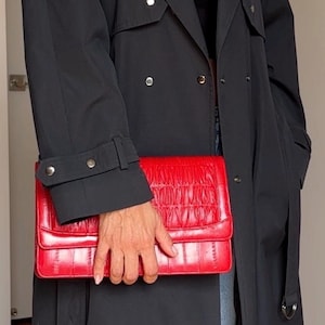 May include: A black trench coat with silver-toned buttons and a belted cuff, paired with a rectangular red clutch bag featuring a horizontal pleated texture on the flap and a smooth finish on the lower section, held in a horizontal position.