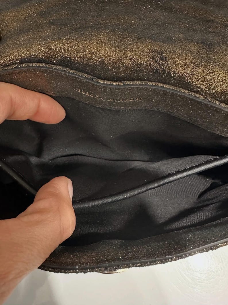 May include: Close-up of a black and gold metallic handbag interior. The bag's interior is black, with a visible pocket and a textured, shimmering exterior. The bag appears to be made of leather or a similar material.