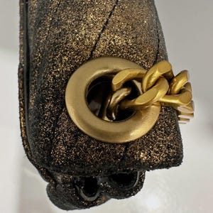 May include: Close-up of a gold and black handbag. The bag has a quilted pattern and a gold chain strap. The hardware is gold-toned. The bag has a sparkly, glittery finish.