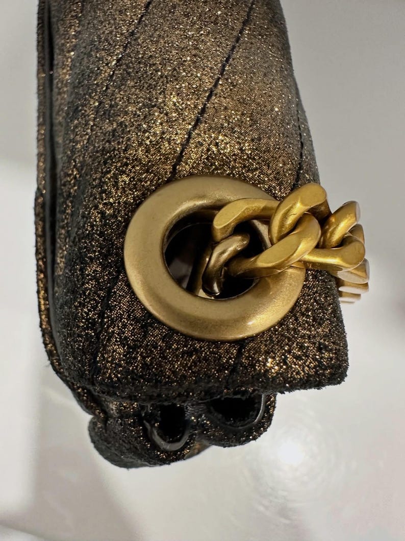 May include: Close-up of a gold and black handbag. The bag has a quilted pattern and a gold chain strap. The hardware is gold-toned. The bag has a sparkly, glittery finish.