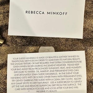 May include: Two white cards with black text on a textured gold background. The top card reads "REBECCA MINKOFF." The bottom card provides care instructions for a suede handbag, including avoiding sunlight, heat, and humidity.