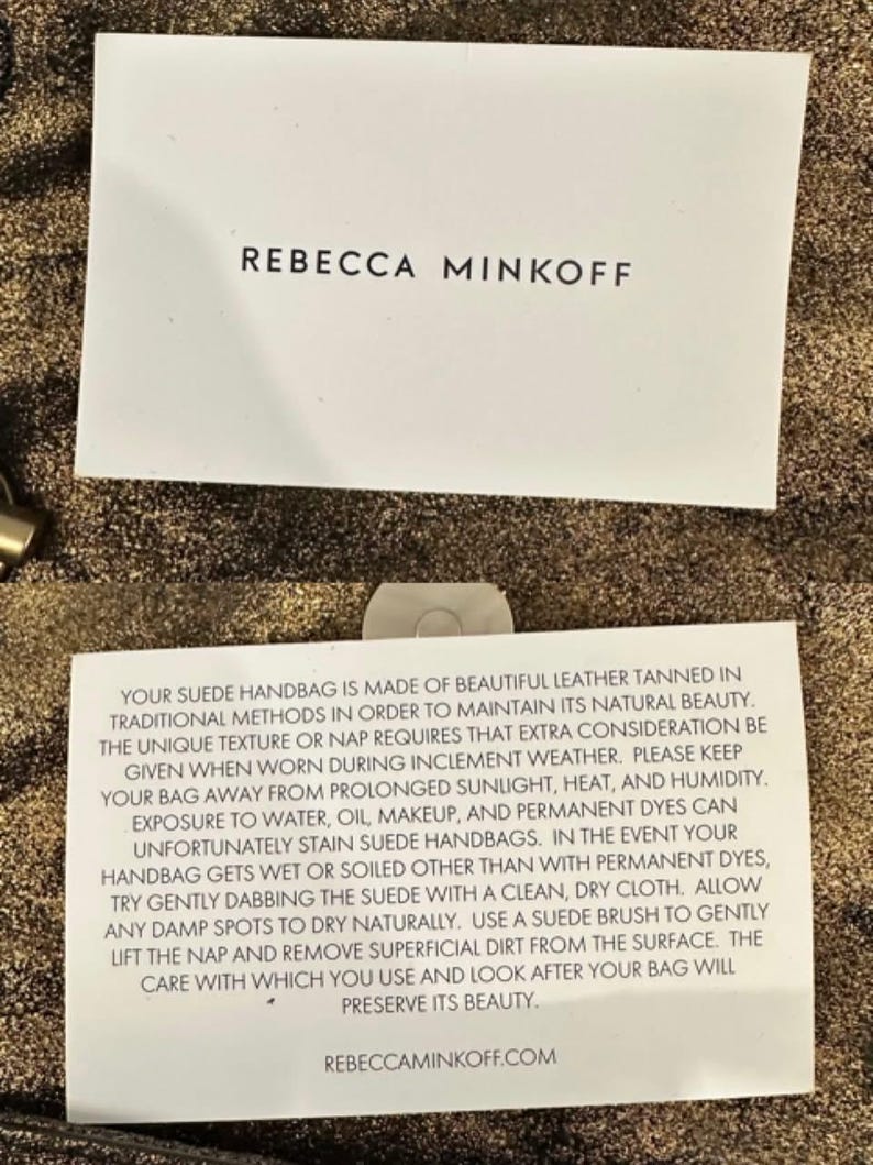 May include: Two white cards with black text on a textured gold background. The top card reads "REBECCA MINKOFF." The bottom card provides care instructions for a suede handbag, including avoiding sunlight, heat, and humidity.