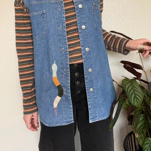 May include: A blue denim vest with a white collar and a colorful embroidered snake design. The vest has a button-up front and is worn over a striped long-sleeved shirt.