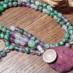 Può includere: Una collana a più fili con perline di pietre preziose verdi, viola e trasparenti. La collana presenta un grande ciondolo rotondo viola scuro e un piccolo ciondolo circolare dorato con un motivo intricato. Le perline sono disposte su una superficie di legno.