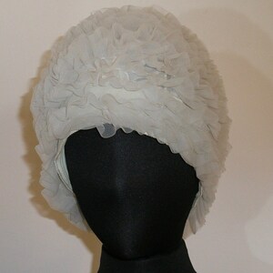 May include: A light gray, vintage-style hat with a ruffled, layered design. The hat is made of a soft, sheer fabric and is displayed on a black mannequin head. The hat's design is reminiscent of the 1950s and 1960s.