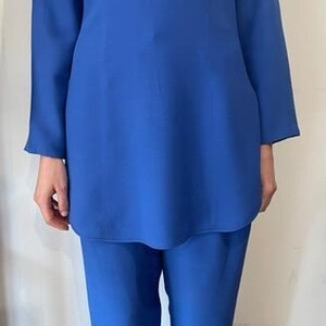 May include: A cobalt blue two-piece suit. The top is a long-sleeved tunic with a high neckline and shoulder pads. The matching pants are straight-legged. The suit is made of a smooth, solid-colored fabric.