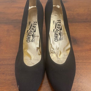 Puede incluir: Un par de zapatos de tacón negros Salvatore Ferragamo. Los zapatos tienen una punta afilada y un tacón alto. El interior del zapato es dorado con el nombre de la marca impreso en negro. Los zapatos están sobre una superficie de madera.