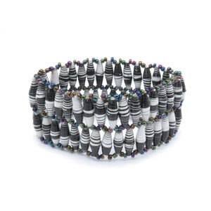 May include: A black and white striped paper bead bracelet with iridescent beads. The bracelet is made of multiple rows of beads and is a handmade piece of jewelry.