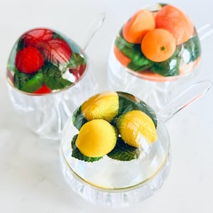 May include: Three clear glass teacups with handles, each filled with different fruit arrangements. One cup has red berries and green leaves, another has oranges, and the third has lemons. The fruit is encased in a clear, dome-shaped cover.