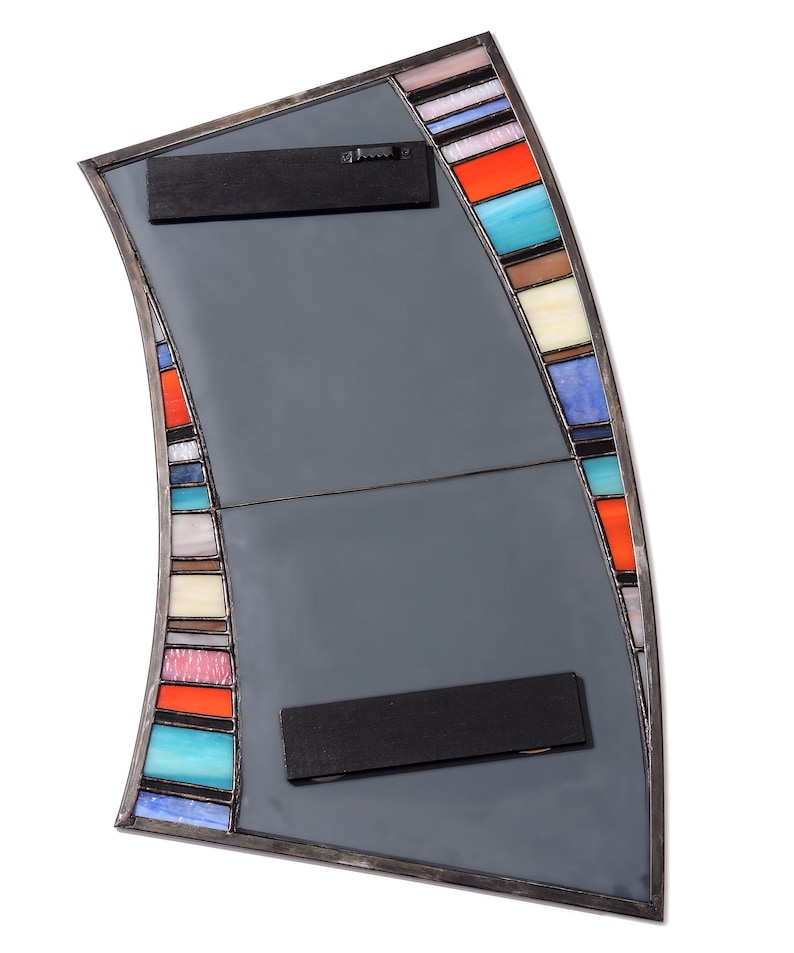 May include: A curved, gray mirror with a stained glass border. The border features colorful stripes in shades of blue, red, orange, yellow, and white. The mirror has two black metal hanging brackets on the back.