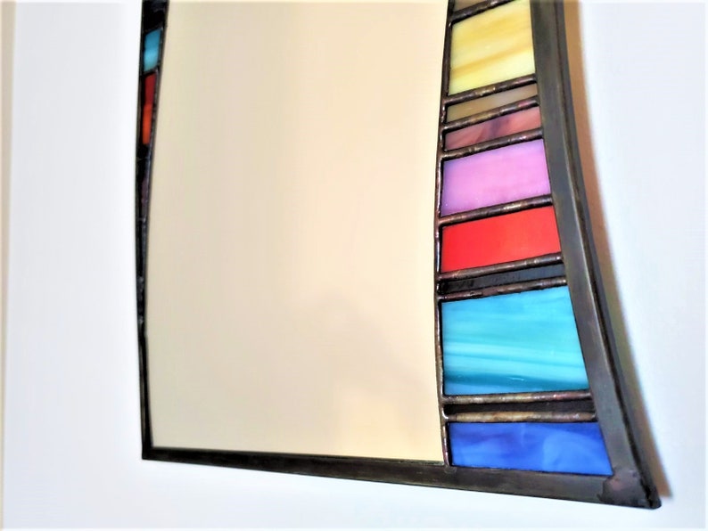 May include: A curved mirror with a stained glass frame. The frame features a series of colorful glass panels in shades of blue, red, yellow, and pink.