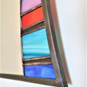 May include: A close-up of a stained glass mirror with a curved frame. The frame is made of metal and has a dark brown finish. The stained glass is in a rainbow pattern, with colors including blue, turquoise, red, pink, and yellow.