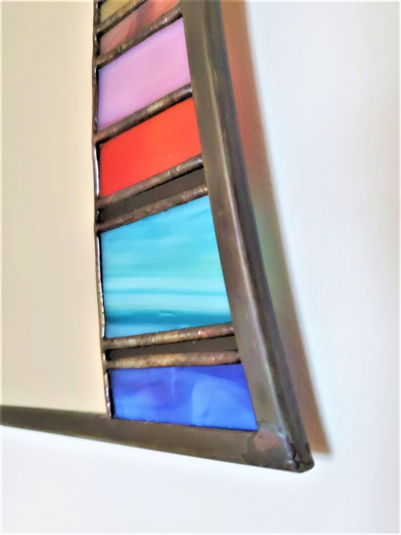 May include: A close-up of a stained glass mirror with a curved frame. The frame is made of metal and has a dark brown finish. The stained glass is in a rainbow pattern, with colors including blue, turquoise, red, pink, and yellow.