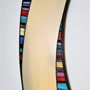 May include: A curved, rectangular mirror with a black frame and a colorful stained glass border. The border features a variety of colors, including blue, red, yellow, and purple.