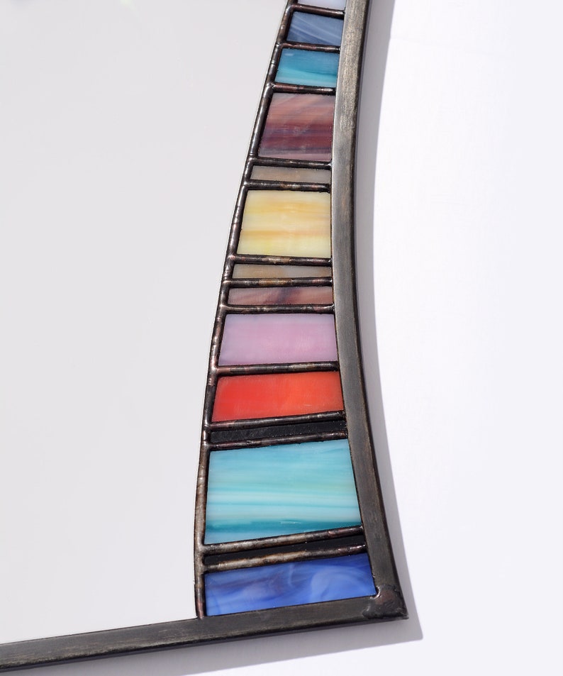 May include: A curved mirror with a stained glass frame. The frame is made of black metal and features a rainbow of colors, including blue, green, yellow, orange, red, and purple.