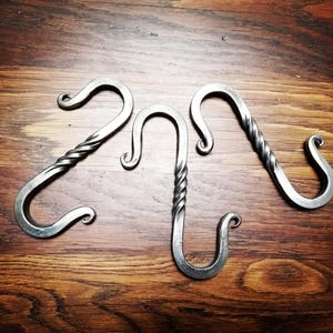 Könnte beinhalten: Drei handgefertigte S-Haken aus Metall mit rustikalem, geschmiedetem Aussehen. Zwei Haken haben ein gedrehtes Design, während der mittlere Haken einen geraden Schaft mit einem gedrehten Abschnitt hat. Die Haken werden auf einer Holzoberfläche präsentiert.