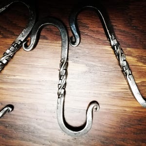 Peut inclure: Trois crochets en S en métal faits à la main avec un design rustique et industriel. Chaque crochet présente un détail en métal torsadé sur la tige et une extrémité recourbée. Les crochets sont présentés sur une surface en bois.