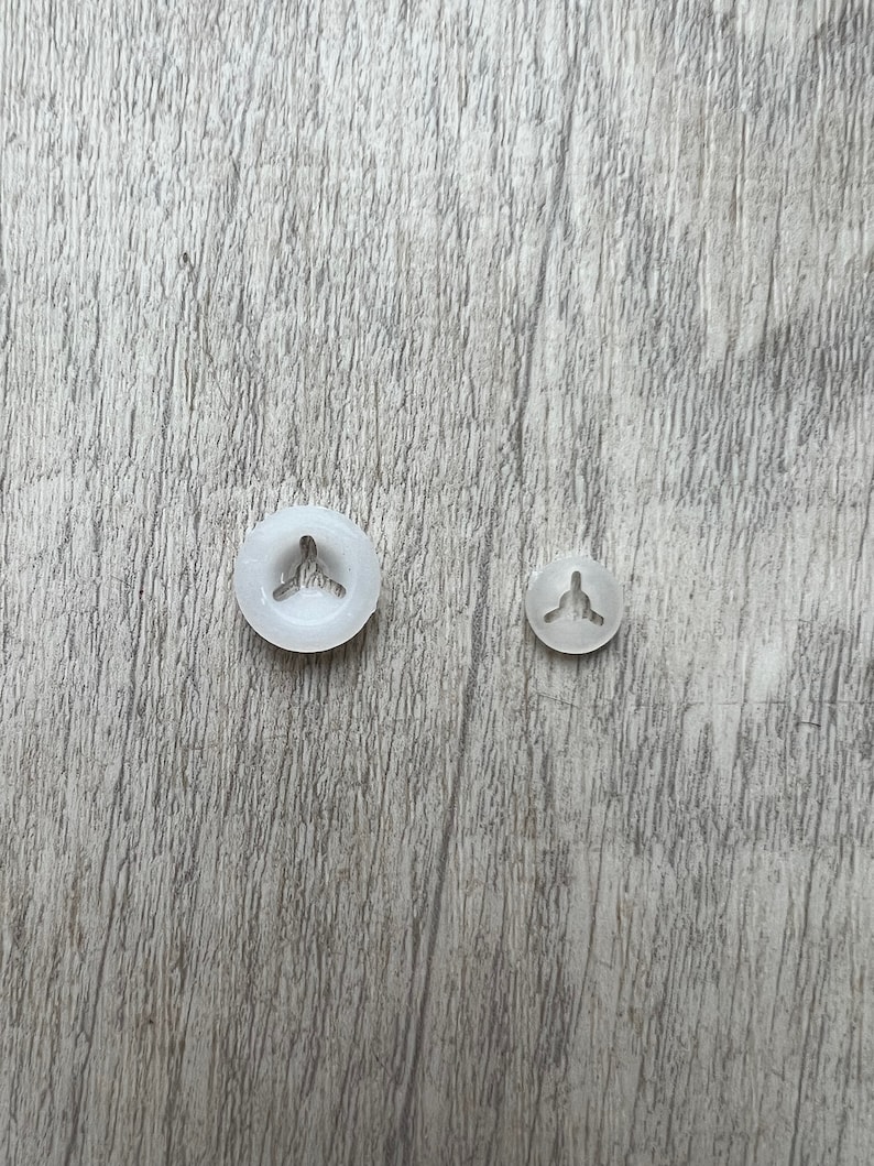 May include: Two white plastic snap fasteners, one larger than the other, on a woodgrain surface.