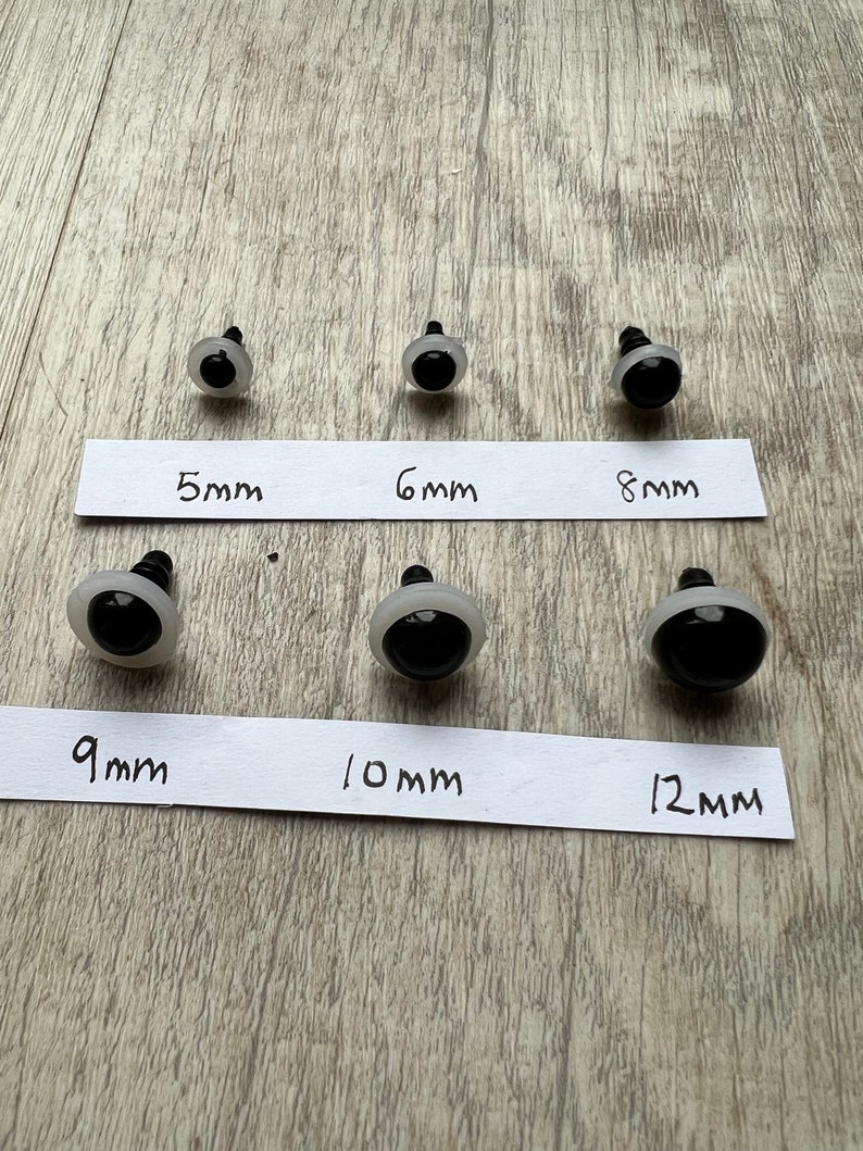 May include: Six pairs of black plastic safety eyes for crafts, ranging in size from 5mm to 12mm. The eyes are on a white surface with labels indicating the size of each pair.