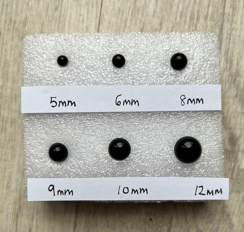 May include: Six black plastic eyes of different sizes, ranging from 5mm to 12mm, displayed on a white foam background. Each eye is labeled with its diameter in millimeters.