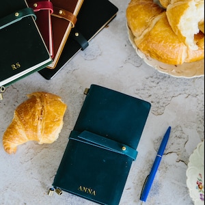 Puede incluir: Tres diarios de cuero verde con letras doradas, uno con el nombre "RS" y otro con el nombre "ANNA". Los diarios están sobre una superficie blanca con dos croissants y un bolígrafo azul.