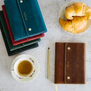 Può includere: Una pila di quattro diari rilegati in pelle di vari colori, tra cui turchese, rosso e nero. Il diario in cima ha il nome "SARAH" in rilievo in lettere dorate. Un solo diario in pelle marrone con il nome "TOM" in rilievo in lettere dorate è mostrato su una superficie bianca. Sono visibili anche una matita e una tazza di tè.