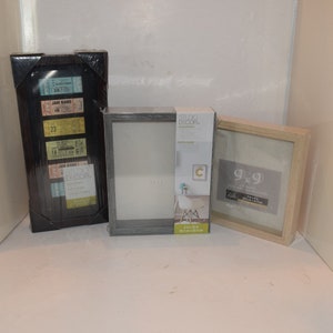 Può includere: Tre cornici per foto, una nera con una collezione di biglietti da concerto, una grigia con un passepartout bianco e una in legno naturale con un passepartout bianco. La cornice grigia ha un'etichetta che dice "Studio Decor" e la cornice in legno ha un'etichetta che dice "9x9".