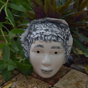May include: A white ceramic planter shaped like a person's head with a textured surface. The planter is filled with green and purple plants.