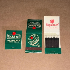 May include: Three matchbooks featuring the Applebee's Neighborhood Grill & Bar logo. Two are closed, displaying a green cover with a red apple icon and white text. One is open, revealing dark brown matchsticks with white tips and branding.
