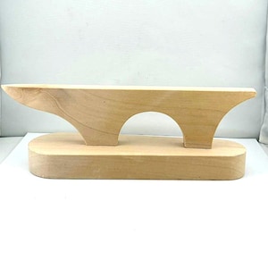 May include: A light-colored wooden tailor's clapper featuring a smooth, rectangular base and a top piece with a curved arch cutout. The tool has a natural wood grain finish and is designed with a tapered, elongated shape for pressing seams.